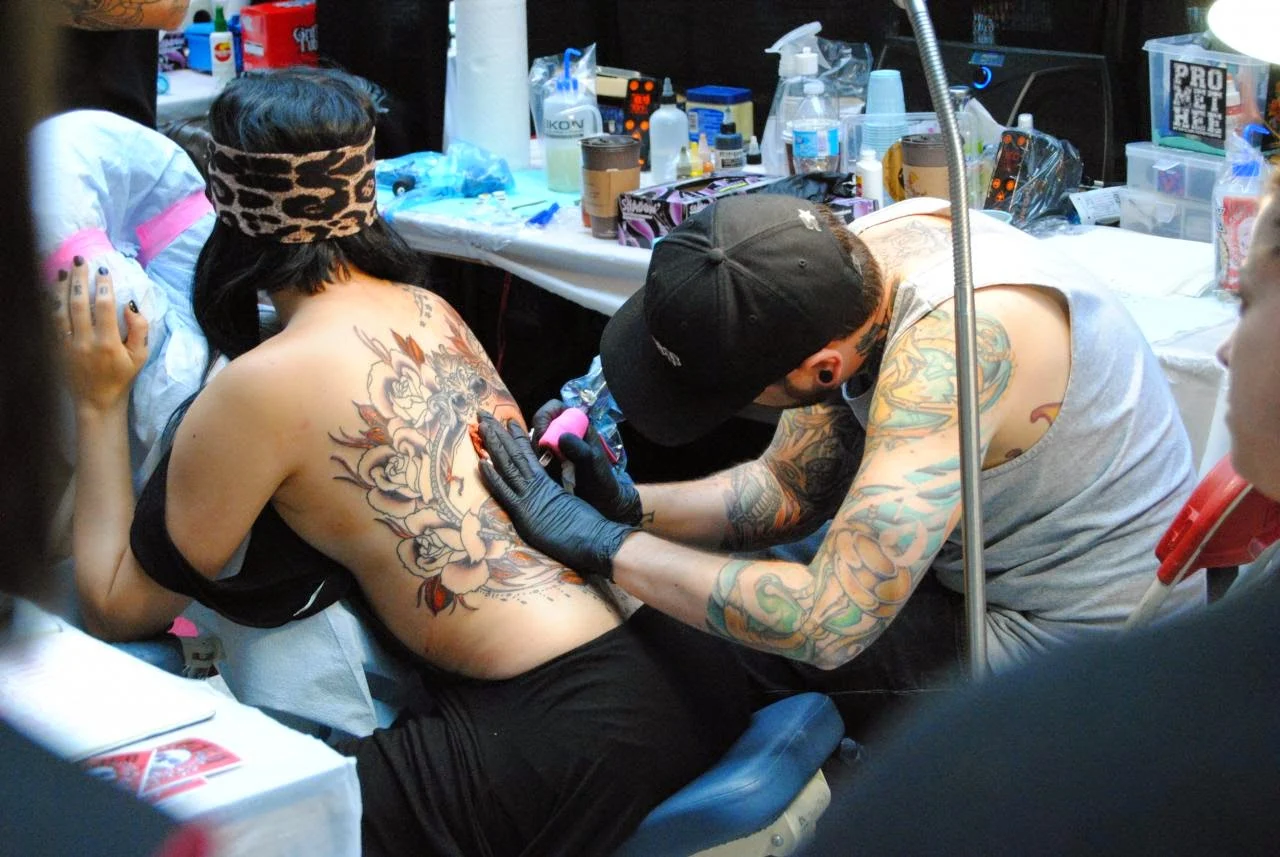 Vemos a una chica de espaldas , y a un tatuador tatuando su espalda con  tatuaje de rosas estilo old school
