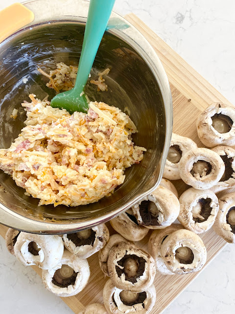 Wooden cutting board with button mushrooms and bowl of stuffed cheese mixture.