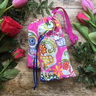 Two drawstring bags made in bright pink fabric with mexican sugar skulls displayed on a wooden surface with pink flowers surrounding