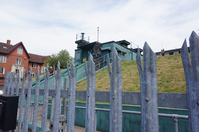 The outside view of Yorks Cold War Bunker, a metal fence with spikes, a bottle green metal building ontop a mound of grass with houses in the background