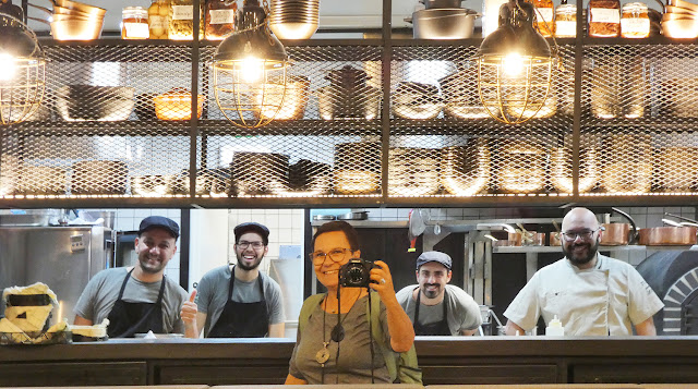 uma mulher com uma máquina fotográfica e 4 homens sorrindo num balcão de cozinha