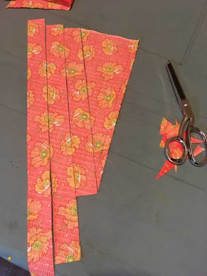 A rough triangle of bright coral fabric with an orange poppy flower print, cut into narrow strips that are barely separated and sitting on a pale blue table with silver sewing shears nearby on top of a pile of very tiny scraps.