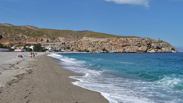 Orilla de playa de arena con el mar con oleaje y acantilados y montañas al fondo.
