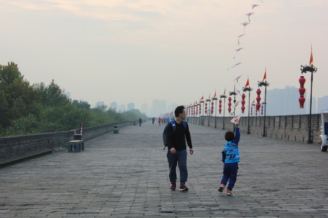 zwiedzanie Xian spacer po murach miejskich, Szerokie pasmo murów z szeregiem lamp z czerwonymi lampionami, w tle majaczące w mgle wieżowce. Po murach idzie ojciec z kilkuletnim dziieckiem, puszczającym latawiec. 