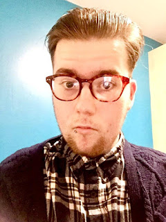 A image taken by myself of myself, a young white male with slicked back dark brown hair with round tortoiseshell glasses a silver lip piercing, a thick black wool cardigan and a grey and white checked shirt on a white background. 