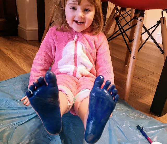 Image of a very happy little girl wearing a pink unicorn onesie and holding her feet in the air. Her feet are covered in blue paint and she is sat on a blue waterproof messy mat.