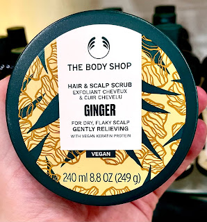 A circular metal silver tin filled with orange scrub with a silver circular tin with a white label that says ginger hair and scalp scrub the body shop in black font on a bright background