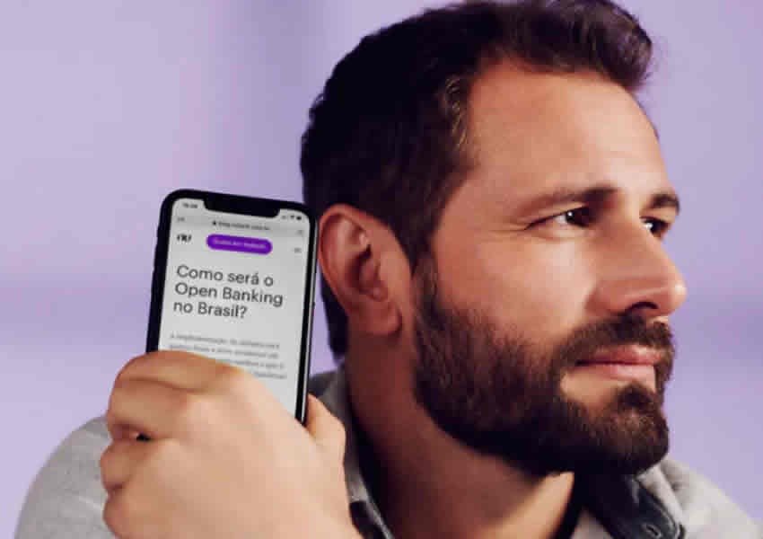Imagem de fundo roxo claro mostra um homem de rosto virado segurando um smartphone com uma das mãos perto do rosto mostrando o app do banco digital nubank.
