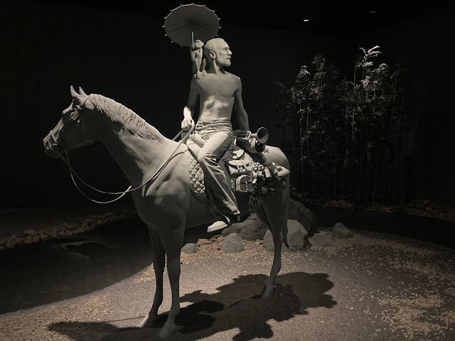 A lifelike but completely grey sculpture of a man in jeans, sitting on a horse, with a monkey on his shoulder..