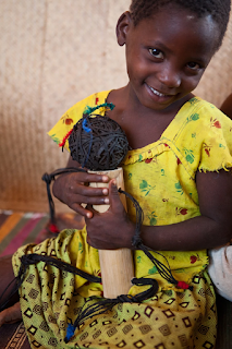 Foto de uma menina negra, moçambicana sentada no chão, cabeça levemente inclinada a direita com leve sorriso nos lábios, segura contra o peito uma boneca feita de bambu e corda preta. Ela tem cabelo bem curto, rosto oval, sobrancelhas retas, olhos que remetem a jabuticabas, nariz largo, lábios grossos e dentes alvos; usa blusa amarela com estampas de flores alternadas em verde e vermelho, saia em tons de amarelo e verde com estampa geométrica marrom. Detalhe da boneca: corpo de bambu descascado, a cabeça é uma bola de corda preta, acima três linhas de lã recobrem três frufrus em verde,vermelho e azul; os braços e pernas da boneca são de meadas de lã em preto com acabamento colorido nas extremidades.