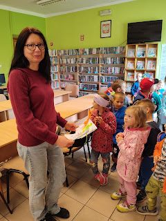 Sala biblioteczna. Pani bibliotekarka trzyma w ręce dwie laurki, które otrzymała od stojącej przed nią grupy przedszkolaków.
