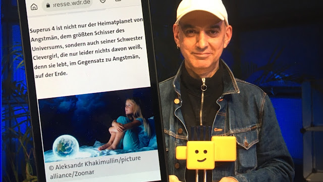 Handy links zeigt Infos zum Hörspiel "Clevergirl", für das Autor Hartmut El Kurdi (rechts im Hintergrund auf einem Pressefoto) den Deutschen Kinderhörspielpreis 2022 bekommen hat