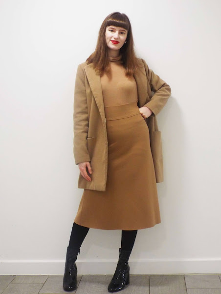 Ellie posed with left hand on left hip, modelling all beige outfit, featuring dramatic, big midi skirt. Paired with beige roll neck, camel smart coat, thick black tights and chunky, high heel, PVC black boots.