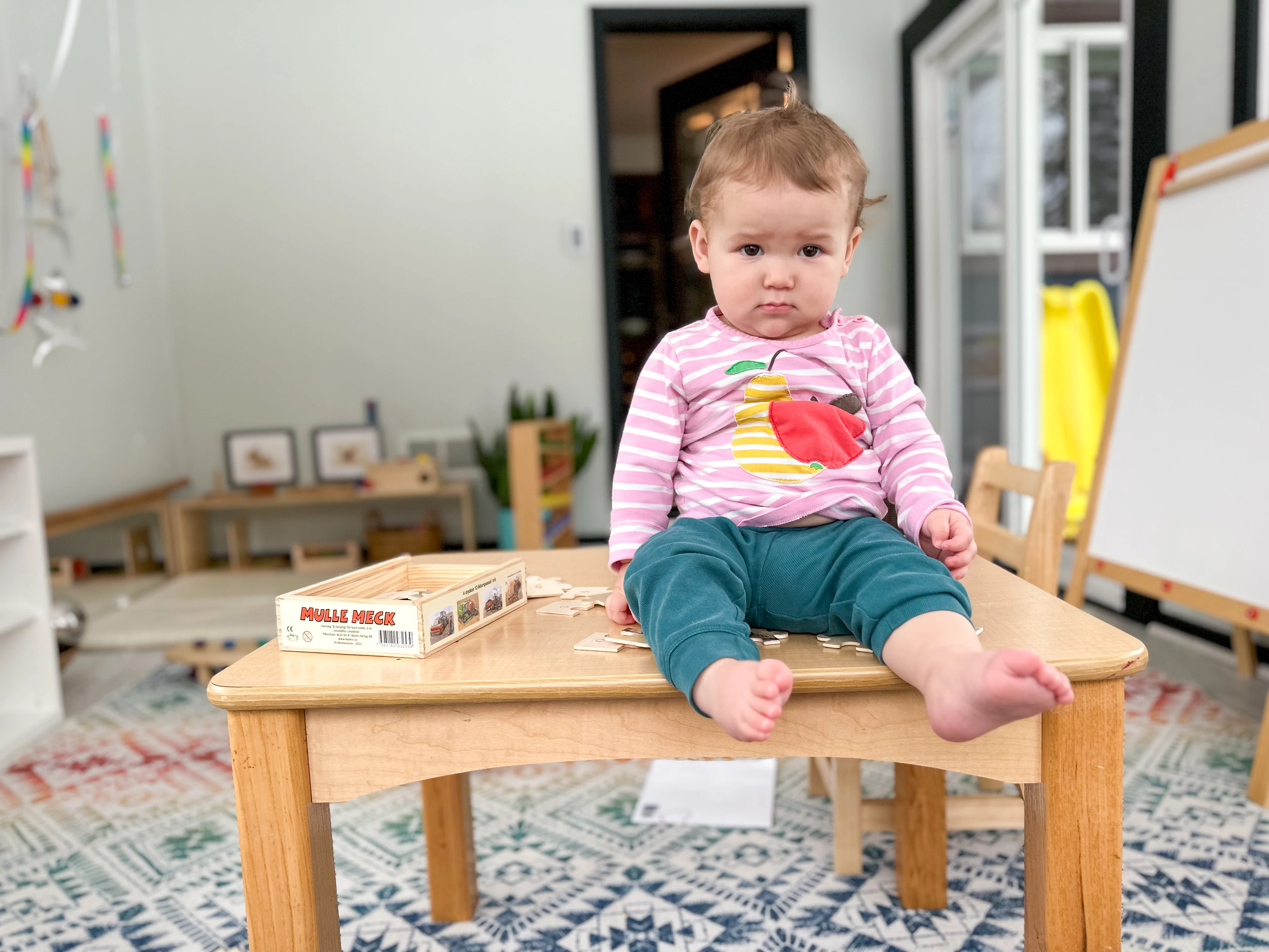 Older Montessori baby sits and looks at camera from on top of a children's table. The background includes her Montessori movement area and playroom.