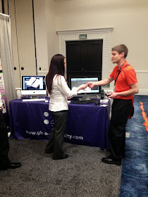 Picture of Derek shaking hands with a young woman in front of a booth with computer screens on its table