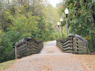 A view as approaching a footbridge in a wooded park. The siderails are worn and there are light-poles to the right.