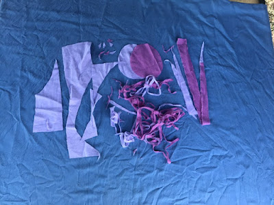 Fabric scraps, in lavender and pink, lie on a blue tablecloth. There are two oval shapes, a couple of larger rectangular-ish pieces, some inch-wide long strips, and a small pile of narrow strips. End ID.