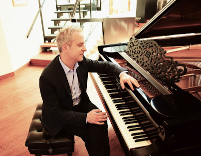 Photo of a white man with gray/white hair, dressed in dark pants and a dark jacket with an open button-down shirt sitting at a piano cross-legged, left arm draped across the piano, looking at the keyboard.