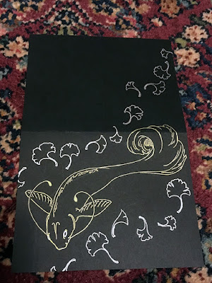 A horizontal pen drawing of a fantastical koi fish between swirling lines of ginkgo leaves on a black greeting card. The fish is in gold, and the leaves are silver.