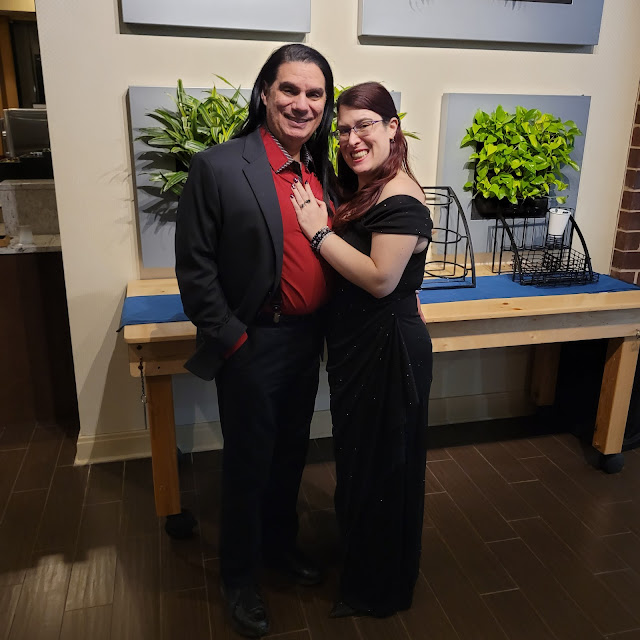 Sarah and Her Husband Ariel at the Sacred Space Gala - He has long black straight black hair, olive skin, and is of regular height. He is wearing a red dress shirt with a black suit. Sarah is short with long red hair and white skin. She is wearing a floor length black sparkly evening gown.