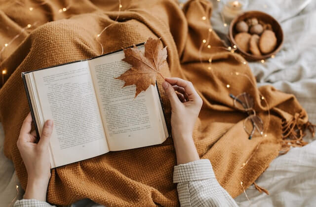 open book on a woman's lap with blanket and autumn leaves