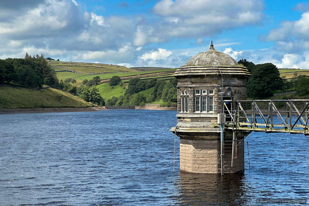 Stonework structure protruding from the blue waters of the reservoir. Green hills behind.