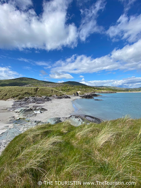 Tall swaying green lush grass on the sand dunes of a white beach with dark grey boulders by a turquoise-coloured ocean under a bright blue sky sprinkled with fluffy clouds.