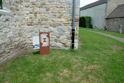 Stèle en béton plantée verticalement dans le sol et peinte avec des fromes colorées aux teintes crayeuses. Un volet en bois est accroché sur la gauche peint avec des formes plus petites.