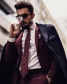 Totally Italian looking dude wearing a burgundy suit and sunglasses and walking down the street smoking a cigar with a long camel hair jacket