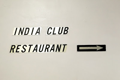 Individual black letters on pale gold backgrounds are stuck onto a cream wall. They say INDIA CLUB RESTAURANT and to their right is stuck a gold arrow on a black background,  pointing right.