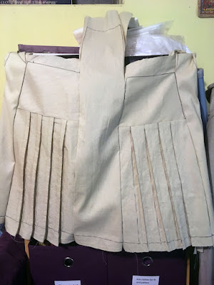 A beige half-skirt propped against purple fabric organizer cubes, with seam lines marked in black sharpie and large seam allowances, showing the rectangular back panels with pleated panels below, and plain panels between and to either side of the pockets. The center plain panel sticks straight up from the waist edge, while the edge plain panels are tucked behind the skirt.