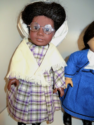 Black doll in pioneer dress of green and purple plaid wearing a shawl, white cap and glasses