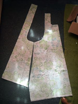 Two pattern pieces cut from patterned green, turquoise, and grey tropical floral fabric, laid out with wrong sides showing. The piece on the left is a tall, slim trapezoid with a long strap protruding from the top edge, aligned with the leftmost edge of the shape. The right piece is almost twice as broad, also roughly trapezoidal, with a slight curve from the edge where it meets the left piece proceeding up to become the left edge of a short strap. A square neckline is cut from the other side of the strap. The hems are mismatched by about five inches, and angled slightly opposite each other to make a ragged down-pointing arrow. Bright sunlight dapples everything.