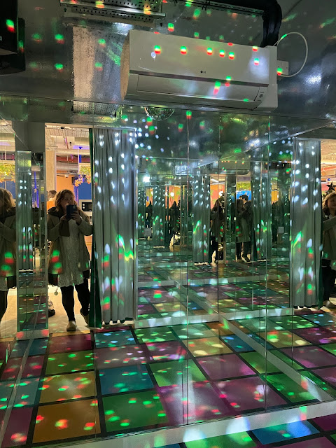 A woman in a white dress stands at the curtained doorway to a small room full of colourful floor tiles and disco lights and takes a photo of herself in the mirrors. The wall is lined with mirrors.