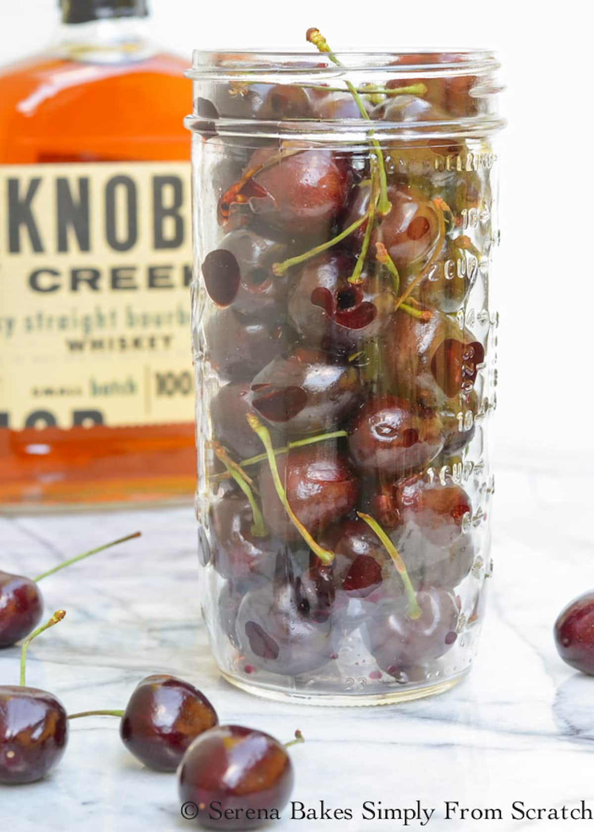 Pitted Cherries in a masons jar with cherries scattered on a white marble counter with a bottle of whiskey in the background to the left side.