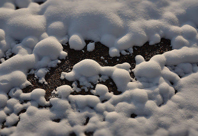 Fluffy snow fell on a driveway of mud and gravel, still above freezing at first, so the snow has melted into lumps and bumps and mounds with dark clay/gravel showing through like tiny valleys in a landscape of ancient worn down mountains