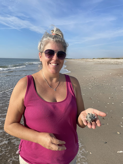 Dad took a picture of me after I found my first conch shell. The tide is going out and is pretty low. The sky is blue and the beach is deserted behind me. I have my head up in a bun with a hot pink tank top on and khaki shorts. I am wearing my sunglasses and am smiling with the conch shell displayed on my open left hand palm.