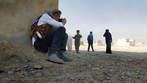 Jan Grarup está sentado no chão, com as castas encostadas em uma muro, fotografando uma criança que vem andando segurando um galho com uma sacola. Logo atrás, vemos uma mulher de burca e um homem andando na direção oposta