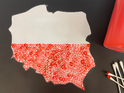 Na czarnym tle przyklejona flaga Polski. Flaga w dolnej części ma czerwone kropki, wykonane farbą. Po prawej stronie leżą: patyczki do wykonywania kropek i butelka czerwonej farby.