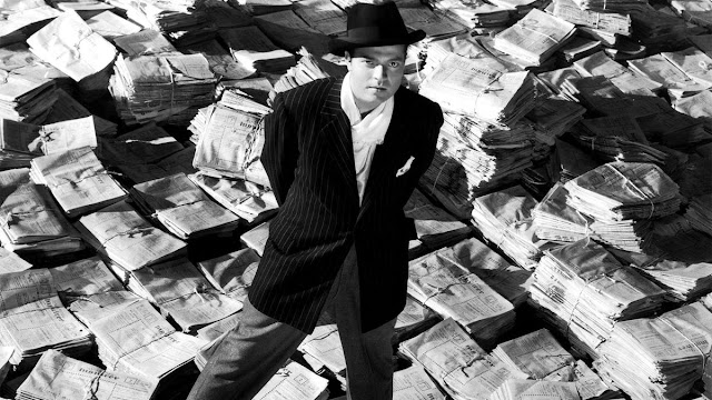 O protagonista Charles Foster Kane está de pé sobre várias pilhas de jornais espalhadas pelo chão. O personagem veste um terno e usa um chapéu. O plano está de baixo para cima e ele olha numa direção próxima à nossa. Podemos notar que ao fundo, à nossa esquerda, está a parte de uma carroça e, do lado direito, um pedaço de um muro de tijolos.