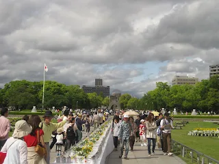Cloudy sky above with trees in the background. Large crowded group of people walking on either side of a very long table going from the foreground to the Atomic Bomb memorial in the background of the photo. The table is covered with candles and flowers.