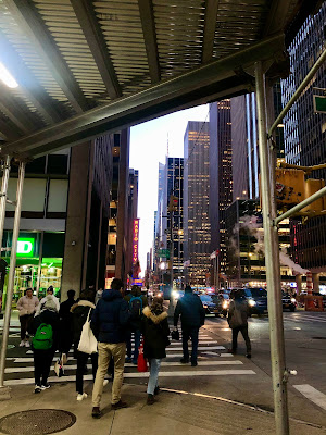 A view of a busy NY sidewalk from under scaffolding, with lights from the buildings including Radio City Music Hall ahead.