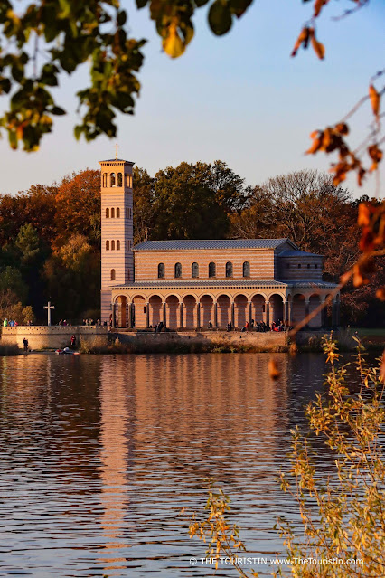 A pastel-coloured church on the banks of a river, reflected in the same, surrounded by autumn foliage trees in the soft light of sunset.