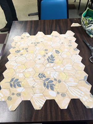 A piece of yellow fabric, wrong-side-up, with a white, pale pink, and black graphic leaf print and edges cut along the outlines of the tesselated hexagons marked over the entire surface, on a dark wood table. A green and turquoise print zippered pouch, small stack of point-to-point half hexagons, and small silver scissors are just visible at upper left.