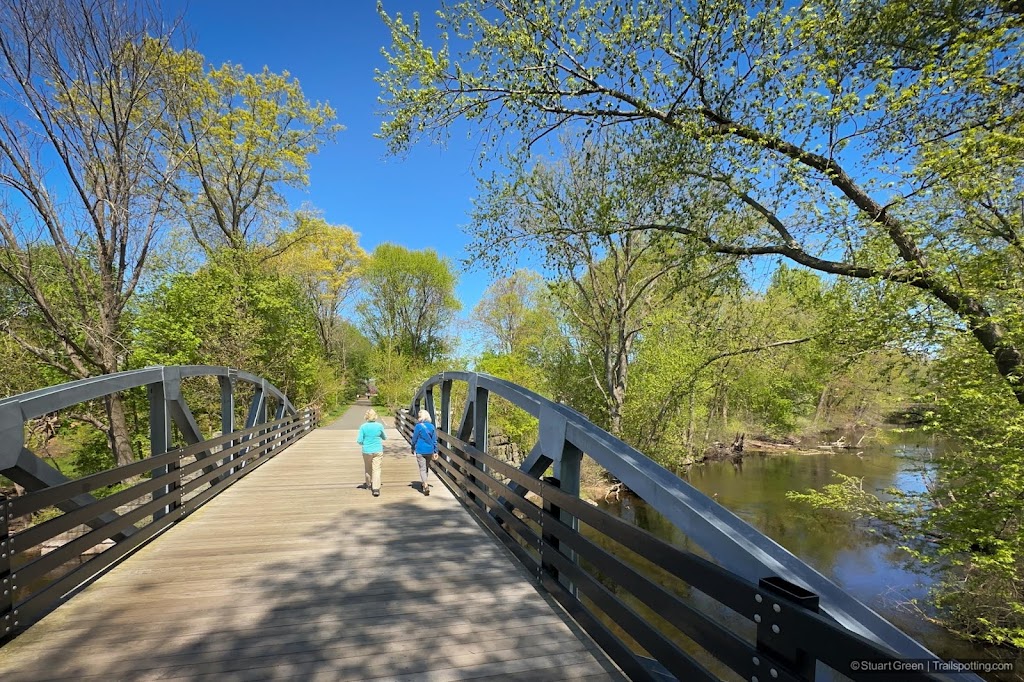 Two people walking across a wooden-decked bridge with steel arch truss supports on the sides. River below with green leafy trees overhanging.