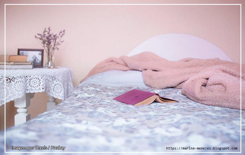 A imagem se trata de uma fotografia colorida, que mostra um quarto, focando em uma cama com lençol branco. Em cima da cama, um livro de capa roxa de cabeça pra baixo. Ao lado da cama é possível ver uma escrivaninha com um porta retrato por cima