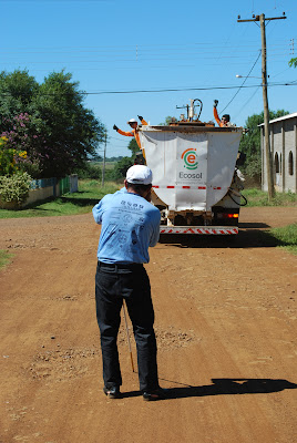 Em pé e de costas para a foto está Luiz apontando a câmera para o caminhão de lixo, que está ao fundo, no caminhão dois homens fazem pose levantando os braços.