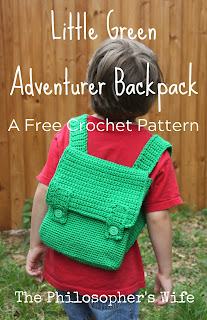 A boy in a red shirt is wearing a crocheted green backpack.  There is wooden fence in the background.  The boy's face is hidden.