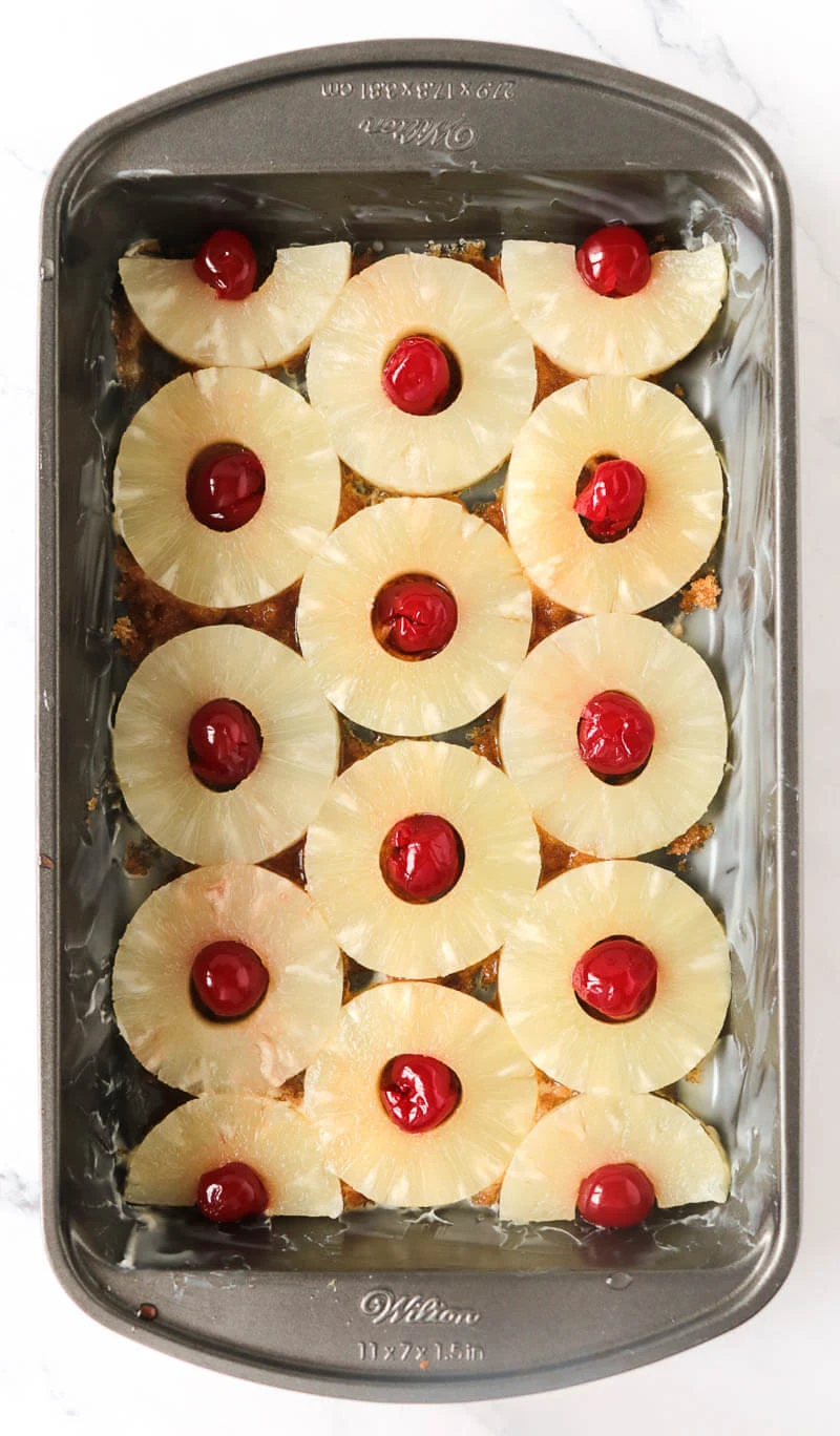 A loaf pan with the base of butter and sugar mixture with pineapples and cherries.