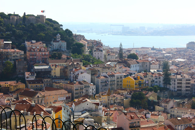 widok Lizbony z tarasu przy kaplicy Nossa Senhora do Monte w pogodny, dzień wakacji. W dole dachy domów o kolorowych fasadach, na nimi wzgórze zamkowe, z wyłaniającymi się między zielonymi koronami drzew wieżami z krenelażami. Na jednej z wież powiewa flaga Portugali. W tle niebieska linia Tagu i nieco zamglony przeciwny brzeg.